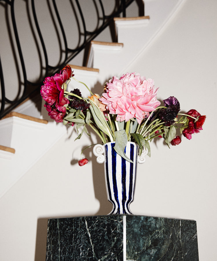The Thallo Pedestal Vase, hand-painted in blue and white and filled with wilted pink and red flowers, stands on a green marble pedestal before a black-railed staircase, making it a striking statement piece.