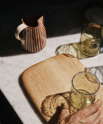 A hand holds a clear glass with a salted rim on a wooden board next to another glass and the Natalia Milk Jug ceramic pitcher on a marble surface.