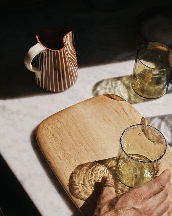 A hand holds a clear glass with a salted rim on a wooden board next to another glass and the Natalia Milk Jug ceramic pitcher on a marble surface.