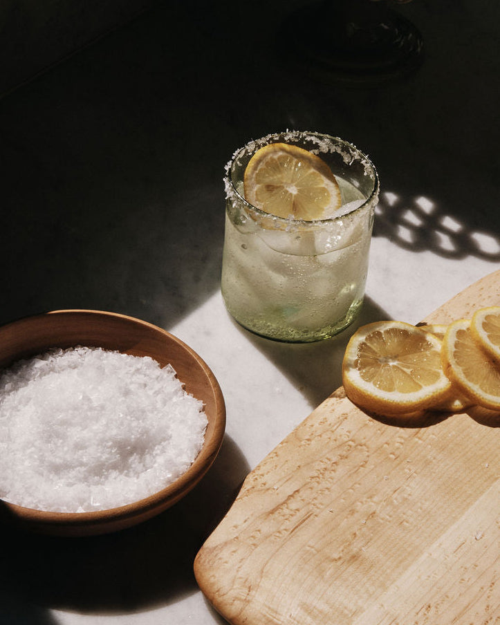 Glass of water with lemon slices, bowl of salt, and cutting board with lemons on a dark surface.