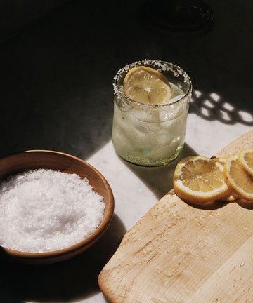 Glass of water with lemon slices, bowl of salt, and cutting board with lemons on a dark surface.
