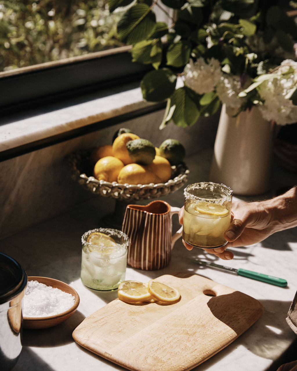 A hand holds a glass of lemonade on a kitchen counter featuring the Birds Eye Maple Serving Board, sliced lemons, a bowl of salt, a ceramic jug, and citrus fruit in sunlight.
