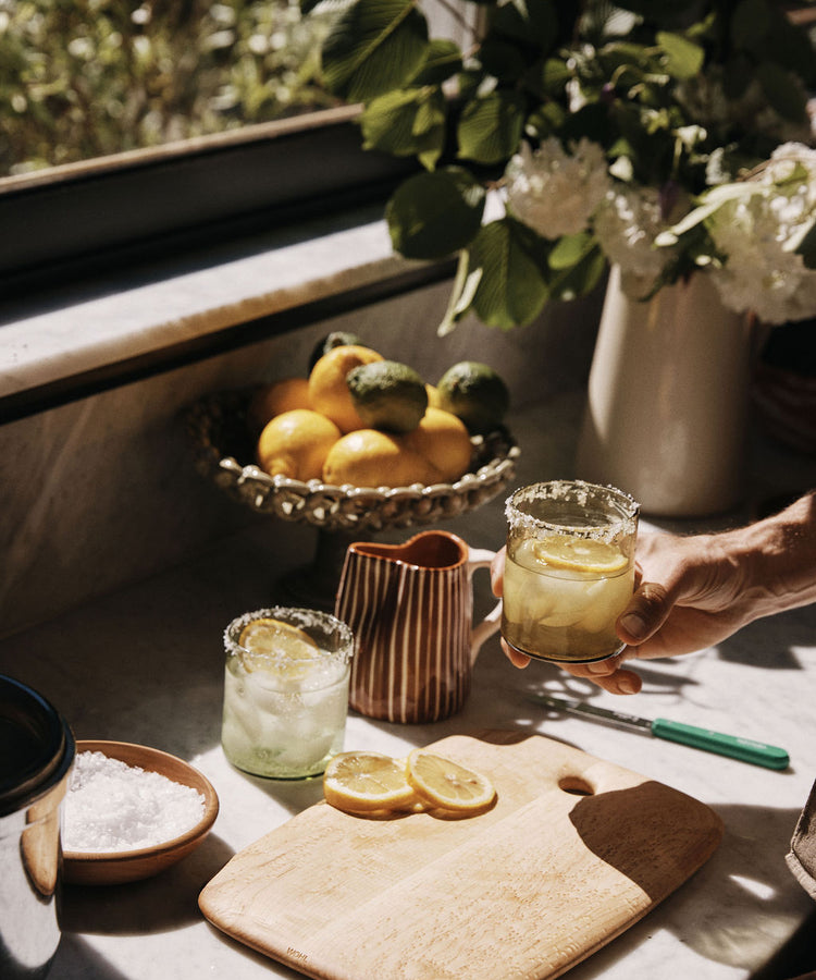 A hand holds a glass of lemonade on a kitchen counter featuring the Birds Eye Maple Serving Board, sliced lemons, a bowl of salt, a ceramic jug, and citrus fruit in sunlight.