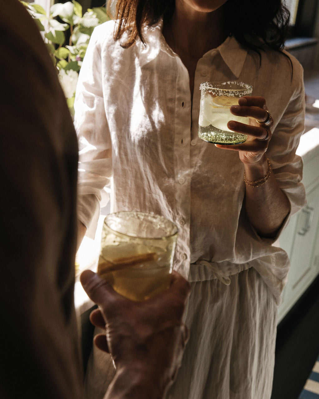 Two people in light clothing hold glasses of a light-colored drink with ice and lemon slices, standing indoors near sunlight. The refreshing beverage is served in the Elodie Straight Glass, Set of 6, adding a touch of artisanal charm to the moment with its elegant handblown design.