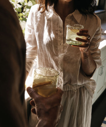 Two people in light clothing hold glasses of a light-colored drink with ice and lemon slices, standing indoors near sunlight. The refreshing beverage is served in the Elodie Straight Glass, Set of 6, adding a touch of artisanal charm to the moment with its elegant handblown design.