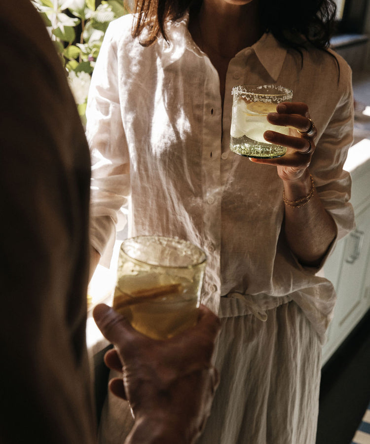 Two people in light clothing hold glasses of a light-colored drink with ice and lemon slices, standing indoors near sunlight. The refreshing beverage is served in the Elodie Straight Glass, Set of 6, adding a touch of artisanal charm to the moment with its elegant handblown design.