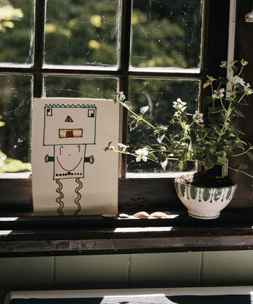 A child's drawing of a robot and a potted flowering plant sit on a sunlit windowsill, beside the Jorge Cereal Bowl (set of 4) featuring a traditional Portuguese pattern, all overlooking lush greenery.