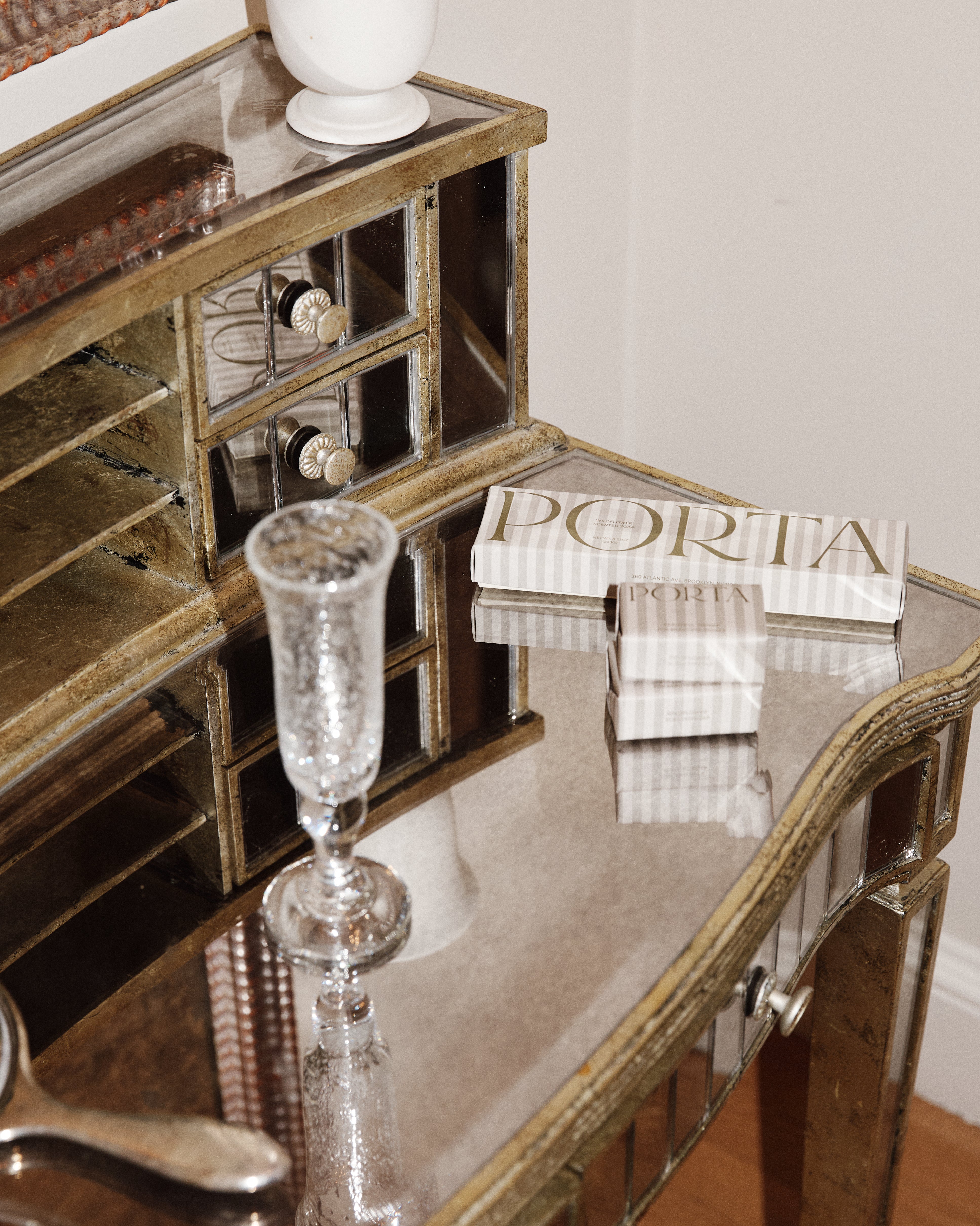 A mirrored vanity table holds a clear glass, a white vase, two books titled PORTIA, and a Porta Soap Box Gift Set with wildflower scent.