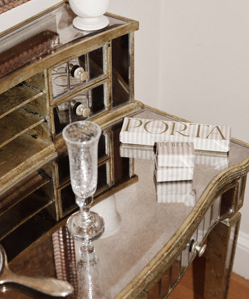 A mirrored vanity table holds a clear glass, a white vase, two books titled PORTIA, and a Porta Soap Box Gift Set with wildflower scent.