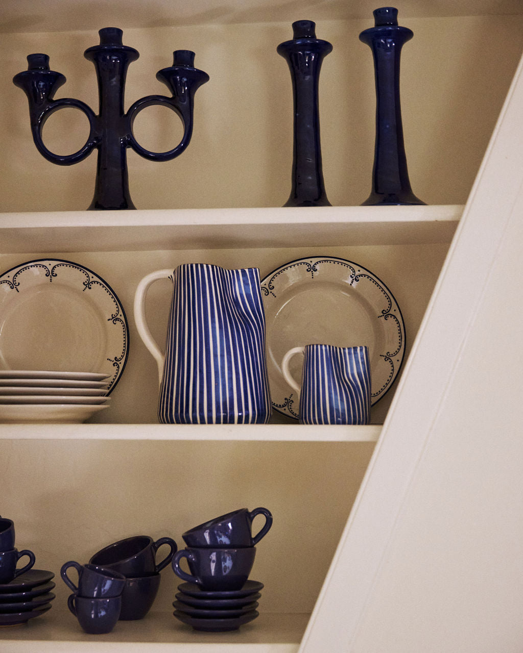 The Natalia Milk Jug, blue candlesticks, and stacked plates are arranged neatly on white shelves.