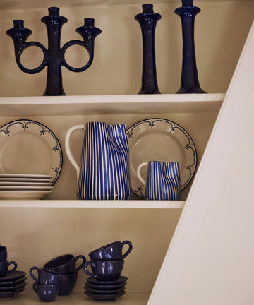 The Natalia Milk Jug, blue candlesticks, and stacked plates are arranged neatly on white shelves.