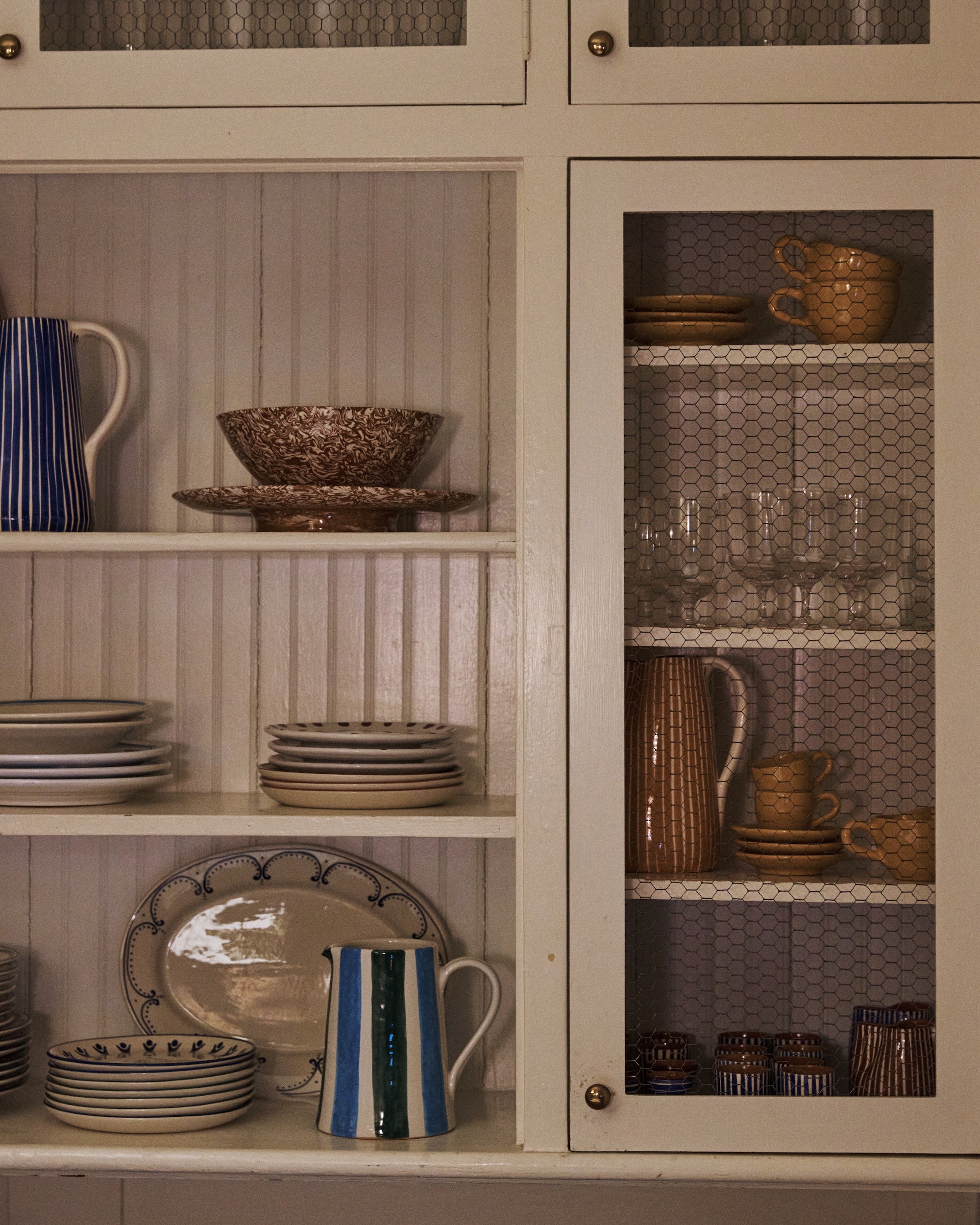 White kitchen cabinet with shelves displaying plates, bowls, mugs, glasses, a striped vase, and the colorful Mina Jug behind chicken wire doors.