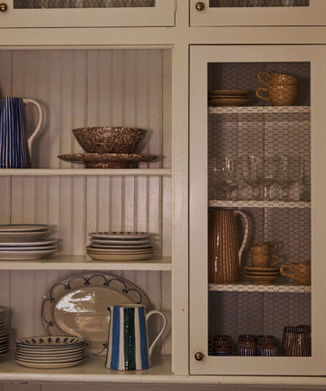 White kitchen cabinet with shelves displaying plates, bowls, mugs, glasses, a striped vase, and the colorful Mina Jug behind chicken wire doors.