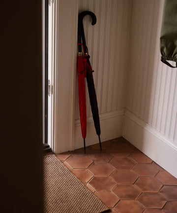 Two closed Victoria Ladies Umbrellas, one red and one black with a leather crook handle, lean against a white paneled wall in the corner of a tiled entryway.