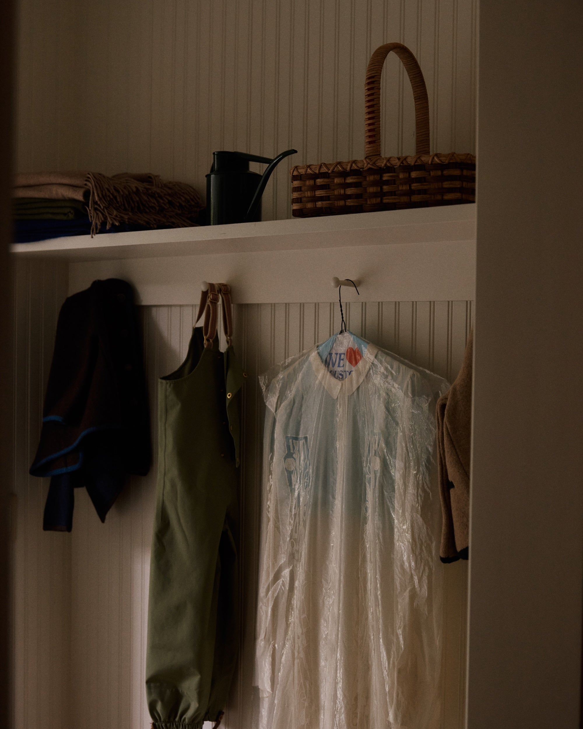 A closet with hanging clothes, a garment bag, a watering can, the Folk Fleur Hand Basket, and folded blankets or coats on a white shelf.