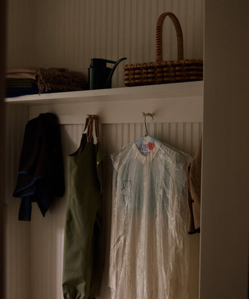 A closet with hanging clothes, a garment bag, a watering can, the Folk Fleur Hand Basket, and folded blankets or coats on a white shelf.