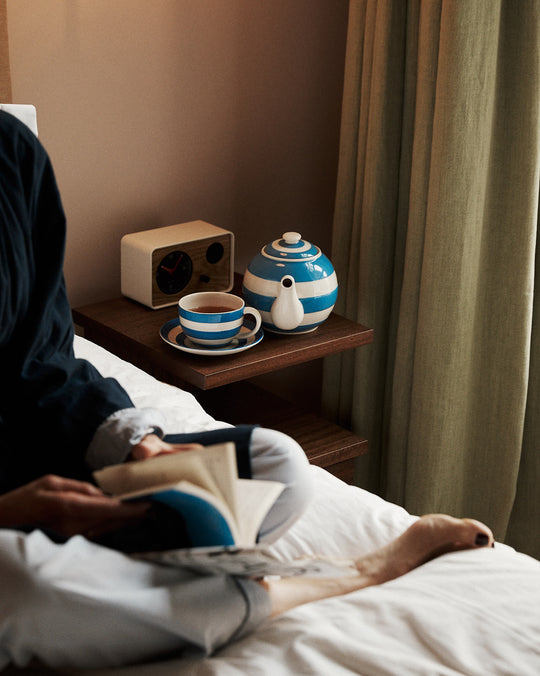 Person lying in bed reading a book with a cup of tea and teapot on a nightstand.