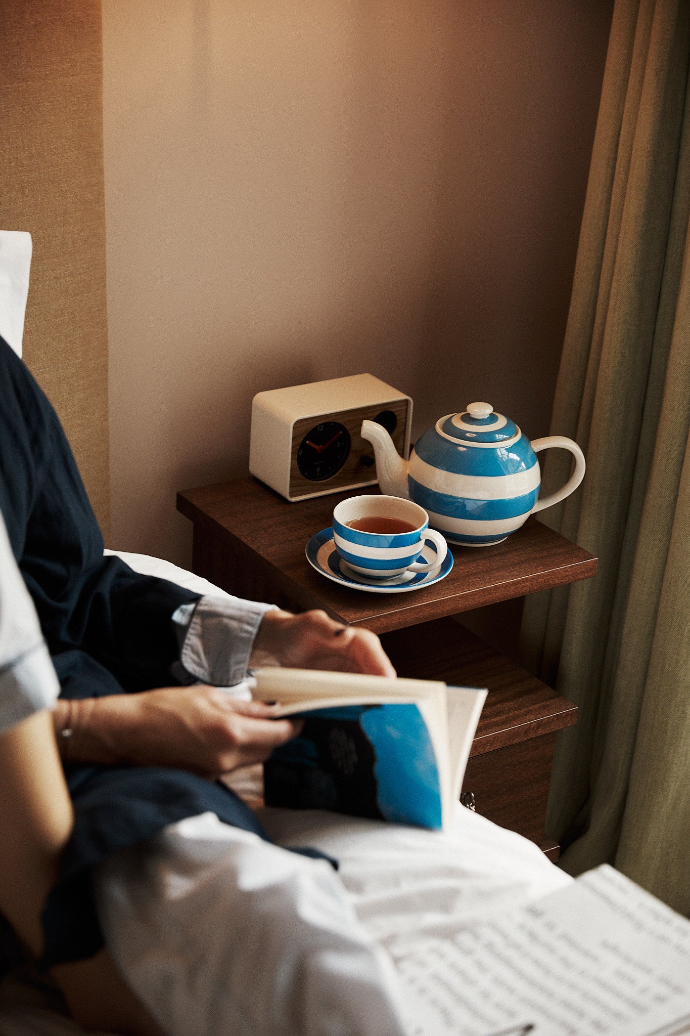 Person reading in bed beside a wooden nightstand with a blue and white striped Betty Breakfast Cup & Saucer Set, matching teapot, and a clock.