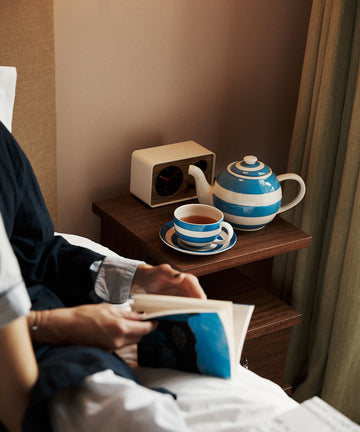 Person reading in bed beside a wooden nightstand with a blue and white striped Betty Breakfast Cup & Saucer Set, matching teapot, and a clock.