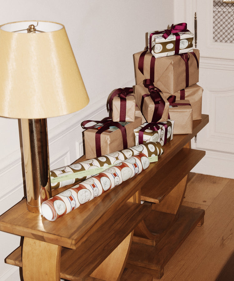 A wooden console table displays gifts wrapped in Intaglio Stripe Gift Wrap with burgundy ribbons, holiday crackers, and a gold lamp, all set against white walls.