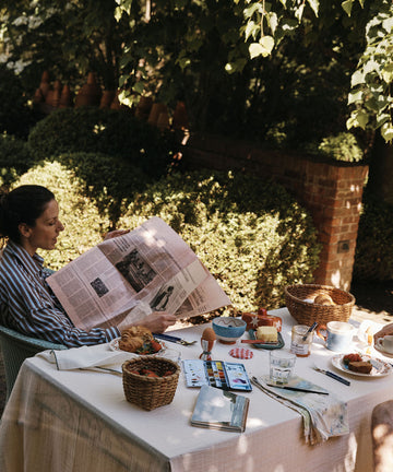 Enjoy a relaxing breakfast outdoors with the Colombe Bundle, featuring cheerful kitchenware and a hand-drawn dove mug from Europe; sunlight filters through trees as you read and use art supplies at the table.