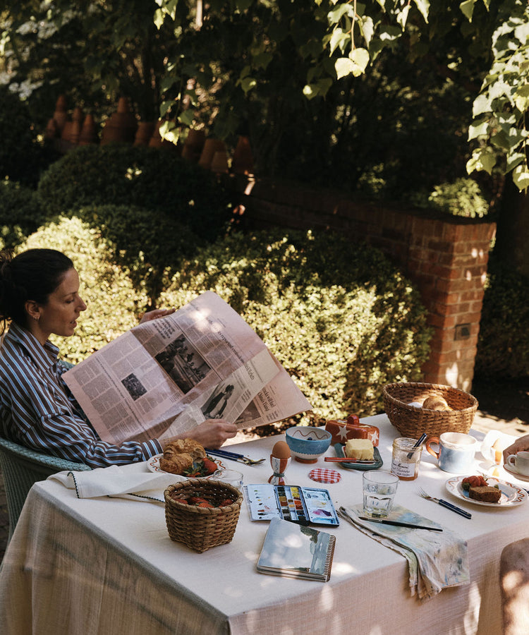 Enjoy a relaxing breakfast outdoors with the Colombe Bundle, featuring cheerful kitchenware and a hand-drawn dove mug from Europe; sunlight filters through trees as you read and use art supplies at the table.