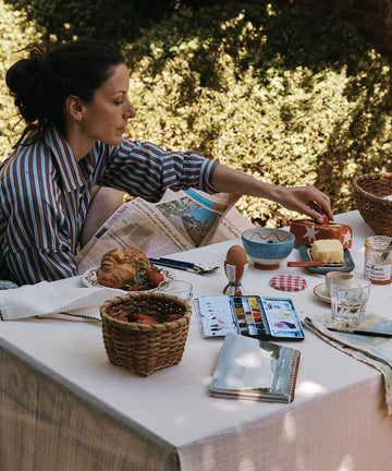 A woman in a striped shirt sits at a table outdoors in a garden setting, serving food from a breakfast spread with a white tablecloth, baskets, plates, and glasses.