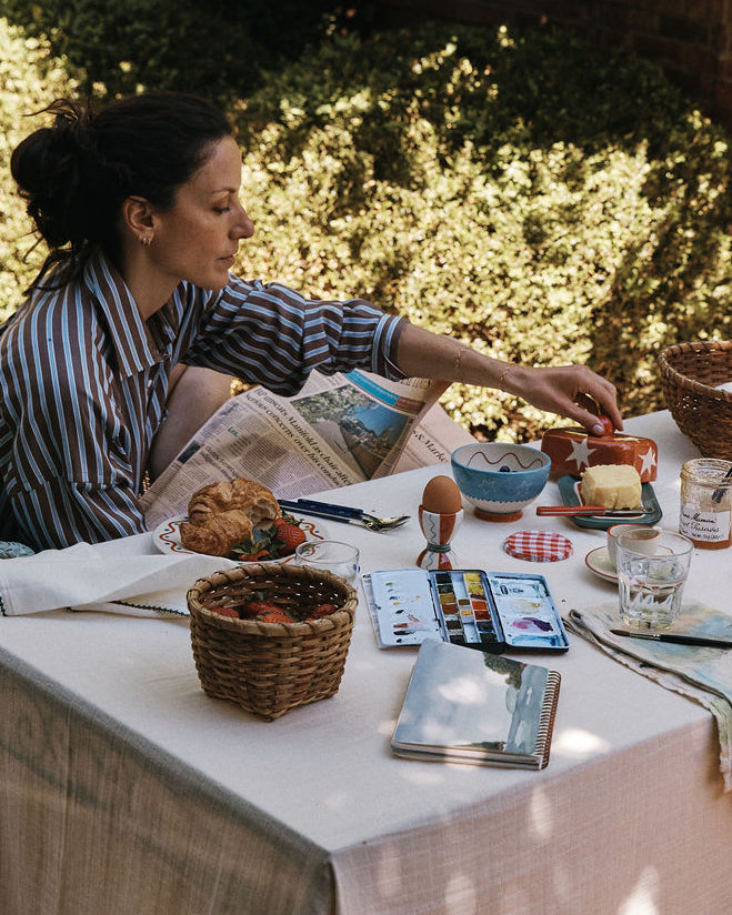 A woman in a striped shirt sits at a table outdoors in a garden setting, serving food from a breakfast spread with a white tablecloth, baskets, plates, and glasses.