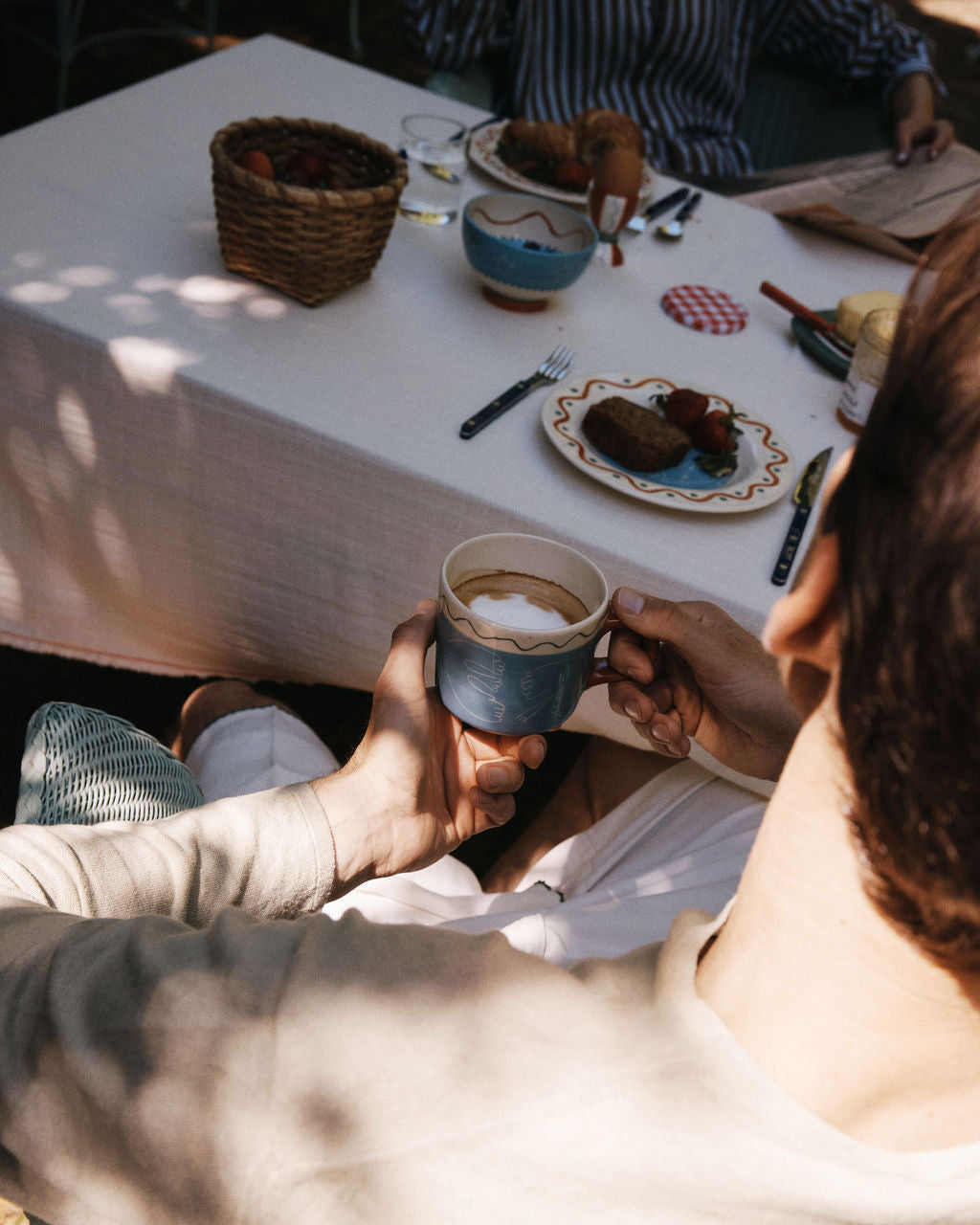 A person holds the Colombe Mug by Laetitia Rouget at an outdoor table adorned with playful tableware, plates of food, bread, and a basket of strawberries in dappled sunlight.