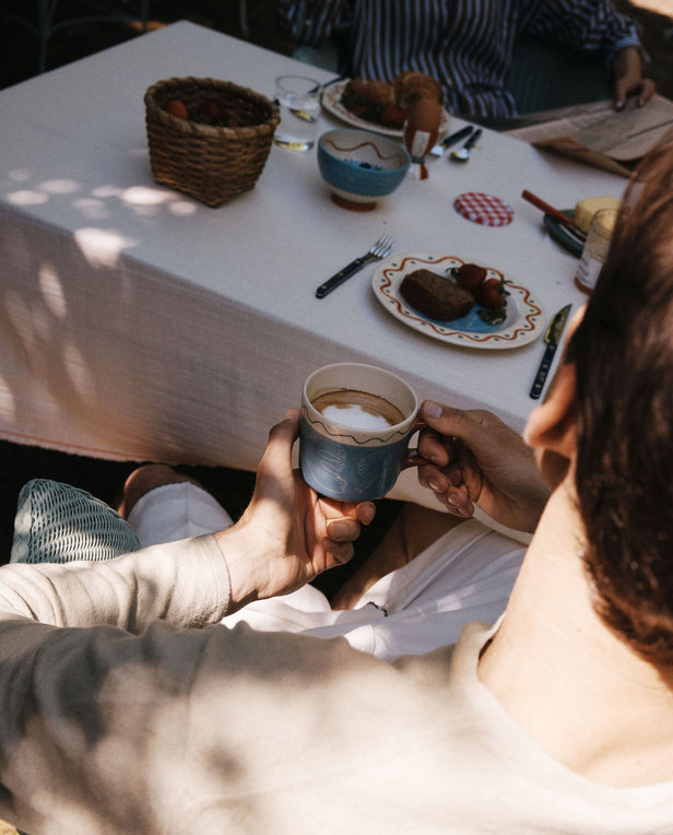 A person holds the Colombe Mug by Laetitia Rouget at an outdoor table adorned with playful tableware, plates of food, bread, and a basket of strawberries in dappled sunlight.