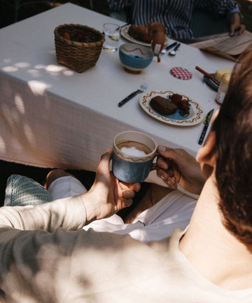 A person holds the Colombe Mug by Laetitia Rouget at an outdoor table adorned with playful tableware, plates of food, bread, and a basket of strawberries in dappled sunlight.