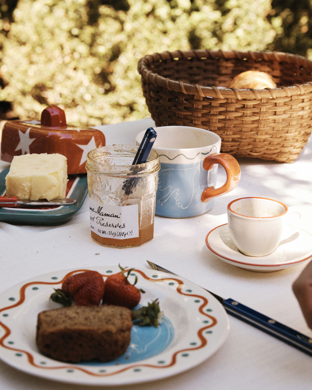 Outdoors on a white tablecloth, the Medium Love Basket holds fresh bread beside strawberries, preserves, butter, mugs, an empty cup, and a knife—ideal for adding rustic charm to your country kitchen decor.