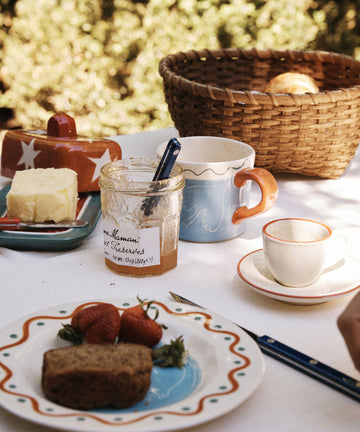 Outdoors on a white tablecloth, the Medium Love Basket holds fresh bread beside strawberries, preserves, butter, mugs, an empty cup, and a knife—ideal for adding rustic charm to your country kitchen decor.