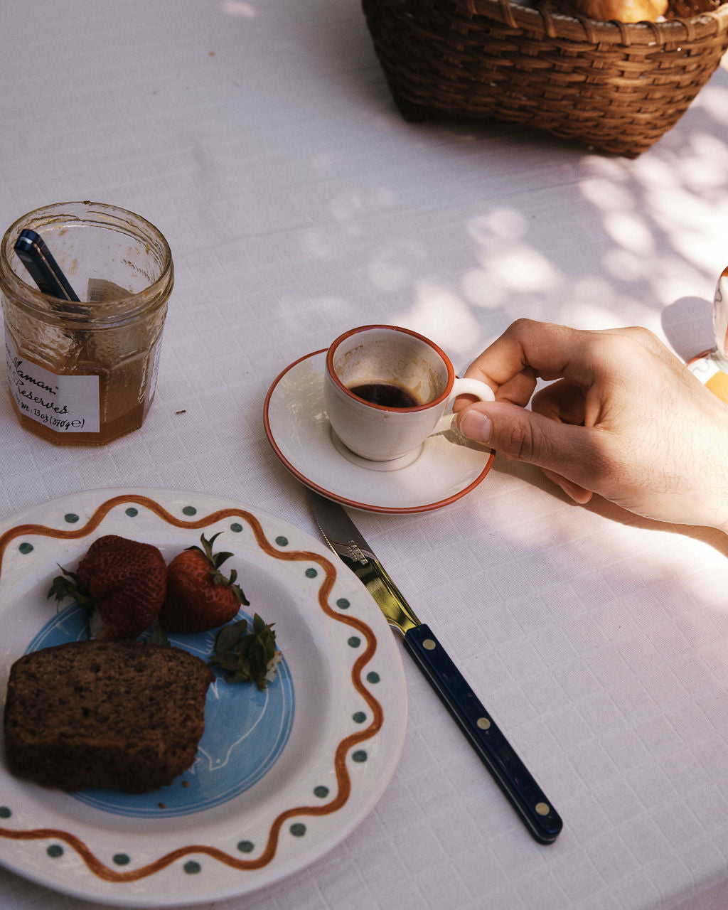 A hand holds the Gio Espresso Set cup on a saucer next to a plate with strawberries, bread, a jar of jam, and a knife atop a white tablecloth.