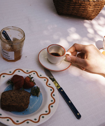 A hand holds the Gio Espresso Set cup on a saucer next to a plate with strawberries, bread, a jar of jam, and a knife atop a white tablecloth.