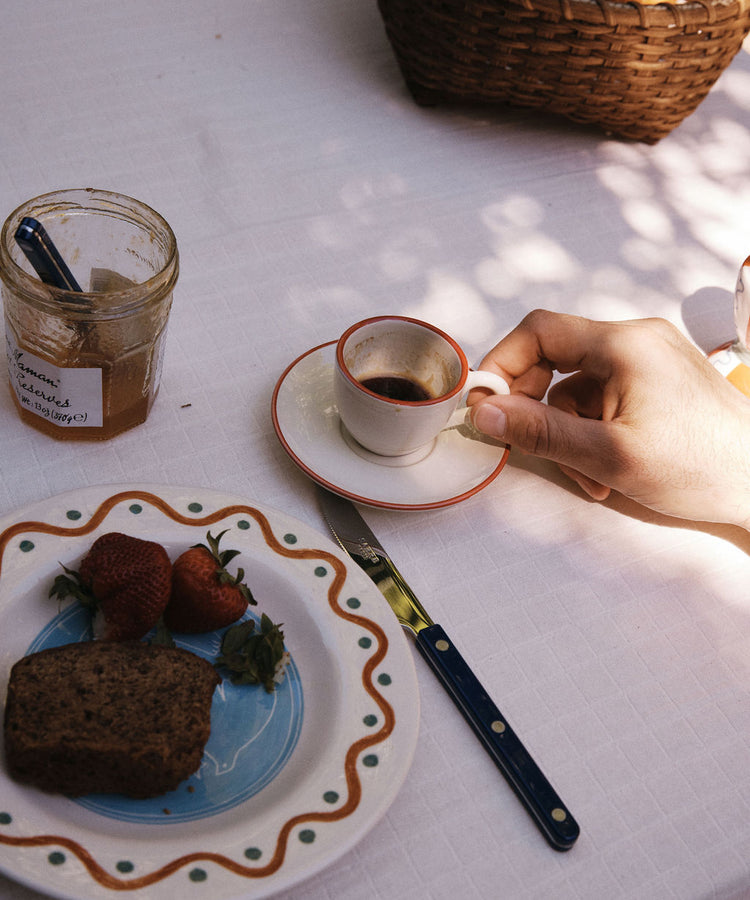 A hand holds the Gio Espresso Set cup on a saucer next to a plate with strawberries, bread, a jar of jam, and a knife atop a white tablecloth.