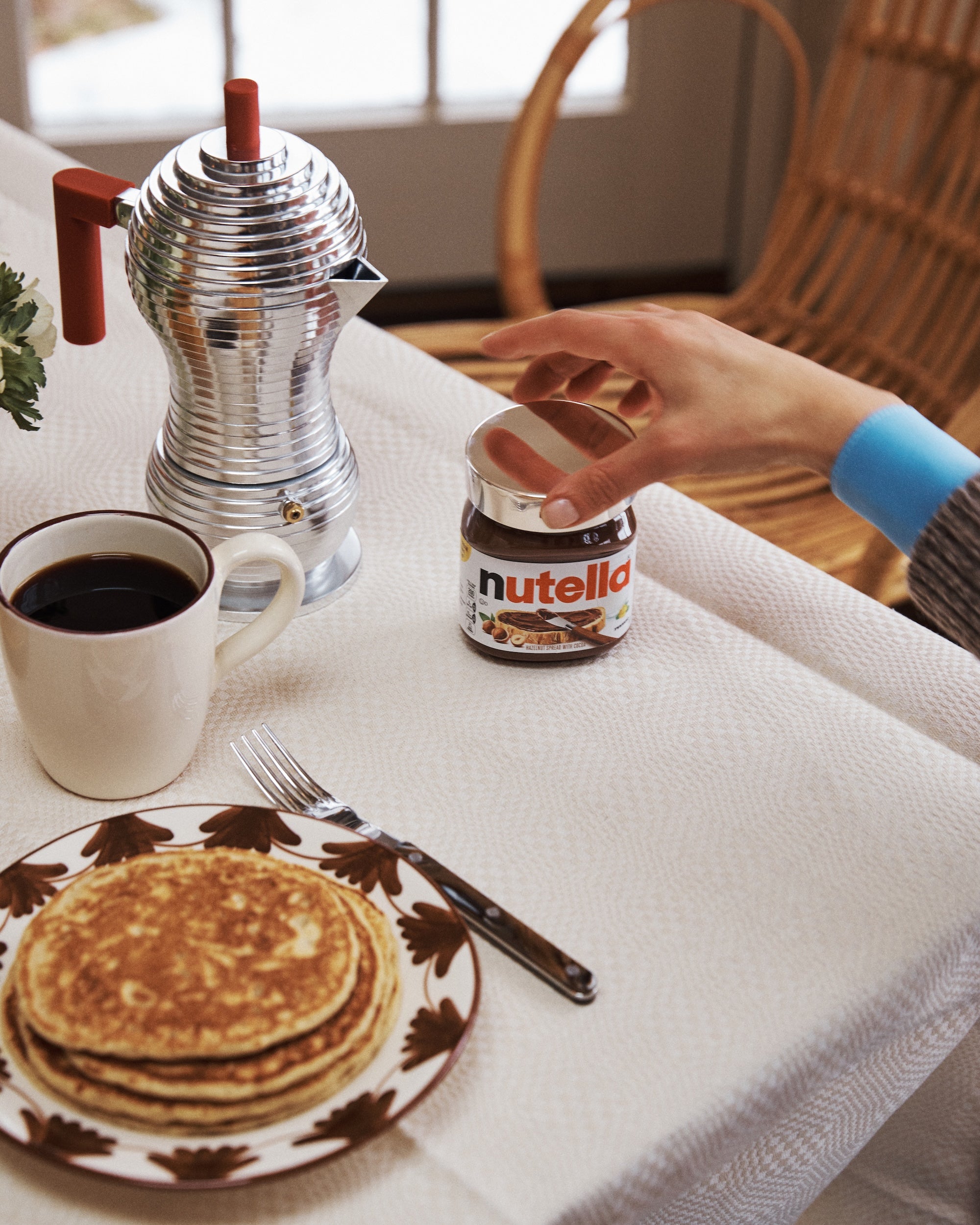 A hand reaches for a jar of Nutella topped with a Sterling Silver Nutella Lid on a breakfast table set with pancakes, coffee, a fork, and an elegant metal coffee pot.