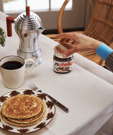 A hand reaches for a jar of Nutella topped with a Sterling Silver Nutella Lid on a breakfast table set with pancakes, coffee, a fork, and an elegant metal coffee pot.