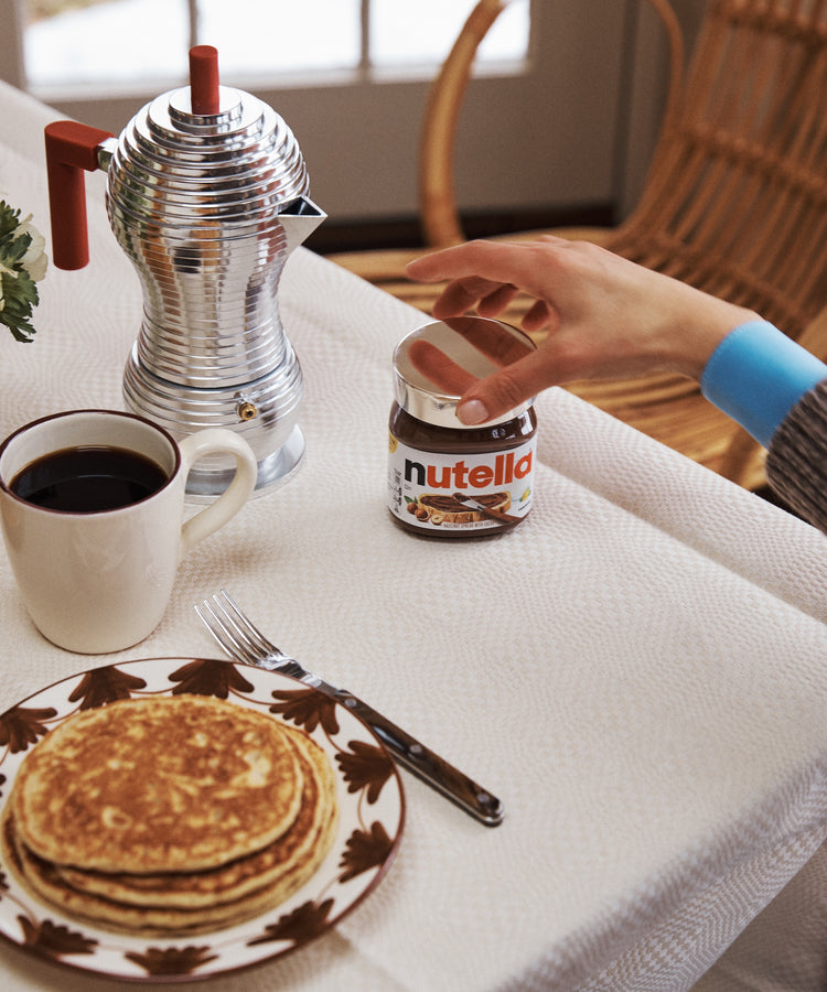 A hand reaches for a jar of Nutella topped with a Sterling Silver Nutella Lid on a breakfast table set with pancakes, coffee, a fork, and an elegant metal coffee pot.