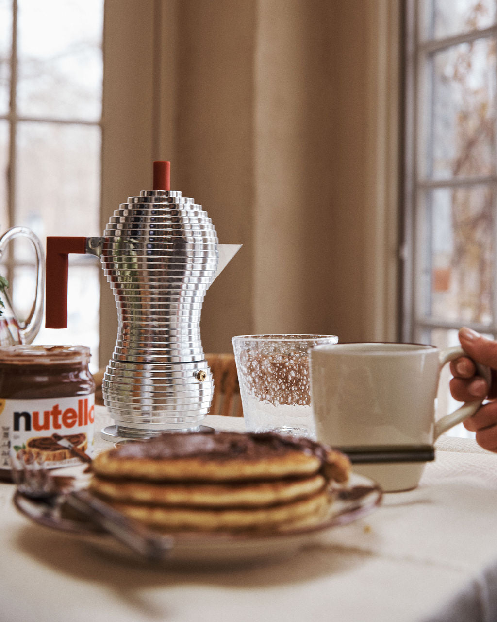 A stack of pancakes, a jar of Nutella, a Pulcina Espresso Coffee Maker, a glass, and a hand holding a mug are on a table near a window.