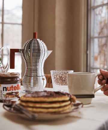 A stack of pancakes, a jar of Nutella, a Pulcina Espresso Coffee Maker, a glass, and a hand holding a mug are on a table near a window.