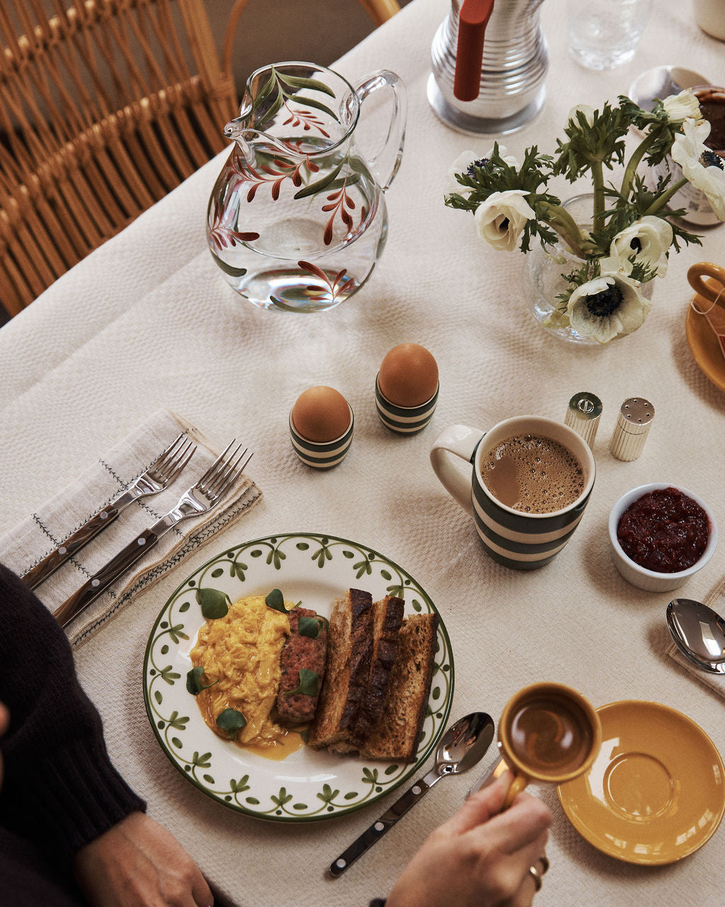 A breakfast table set with scrambled eggs, toast, avocado, soft-boiled eggs, coffee, water in a glass pitcher, jam, and white flowers in a beige setting featuring the unique handmade Botanical Jug.
