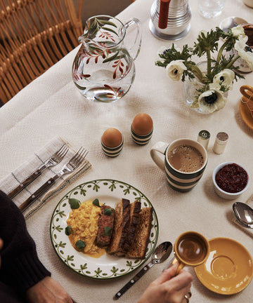 A breakfast table set with scrambled eggs, toast, avocado, soft-boiled eggs, coffee, water in a glass pitcher, jam, and white flowers in a beige setting featuring the unique handmade Botanical Jug.
