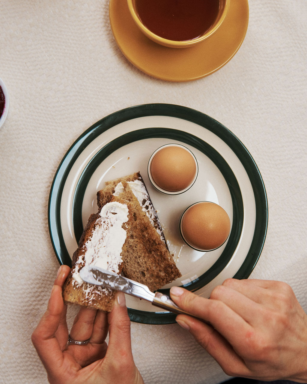 A person spreads cream cheese on brown bread next to two boiled eggs, all served on Betty Breakfast Plates; a cup of tea rests nearby, completing the morning breakfast scene.