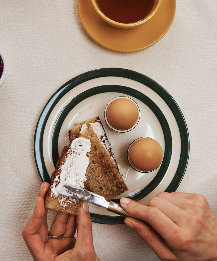 A person spreads cream cheese on brown bread next to two boiled eggs, all served on Betty Breakfast Plates; a cup of tea rests nearby, completing the morning breakfast scene.