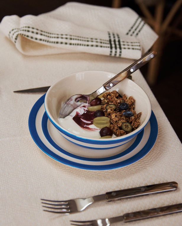A UK West Country pottery cereal bowl with yogurt, granola, blueberries, grapes, and berry sauce sits on a set table featuring a Betty Breakfast Plate, napkin, fork, and knife.