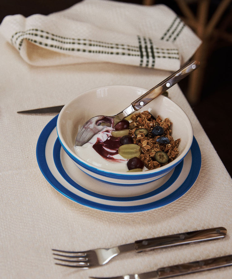 A UK West Country pottery cereal bowl with yogurt, granola, blueberries, grapes, and berry sauce sits on a set table featuring a Betty Breakfast Plate, napkin, fork, and knife.