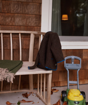 A wooden bench with a green merino wool blanket, an Austrian Wool Jacket, and a toy lawn mower on a porch with fallen leaves.