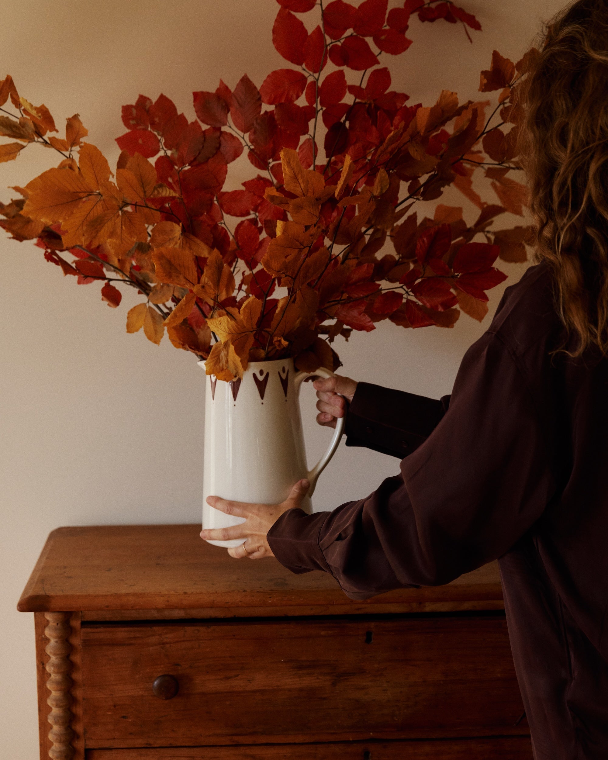 A Vito Jug filled with red and orange autumn leaves sits on a wooden dresser against a plain beige wall.