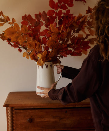 A Vito Jug filled with red and orange autumn leaves sits on a wooden dresser against a plain beige wall.